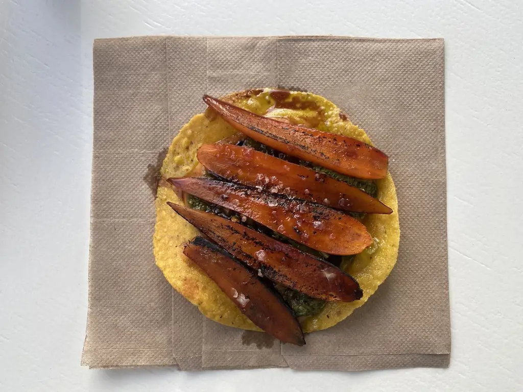 Морковь лежит на золотой тостаде, которая лежит на коричневой квадратной салфетке на белом столе