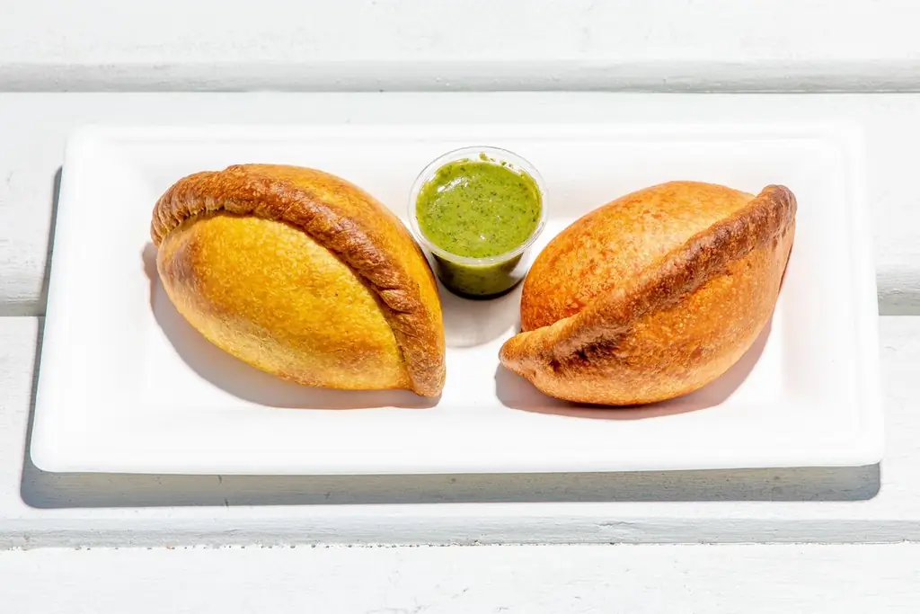 Две золотые сальтены сидят на белой тарелке с контейнером с зеленым соусом льяхуа посередине