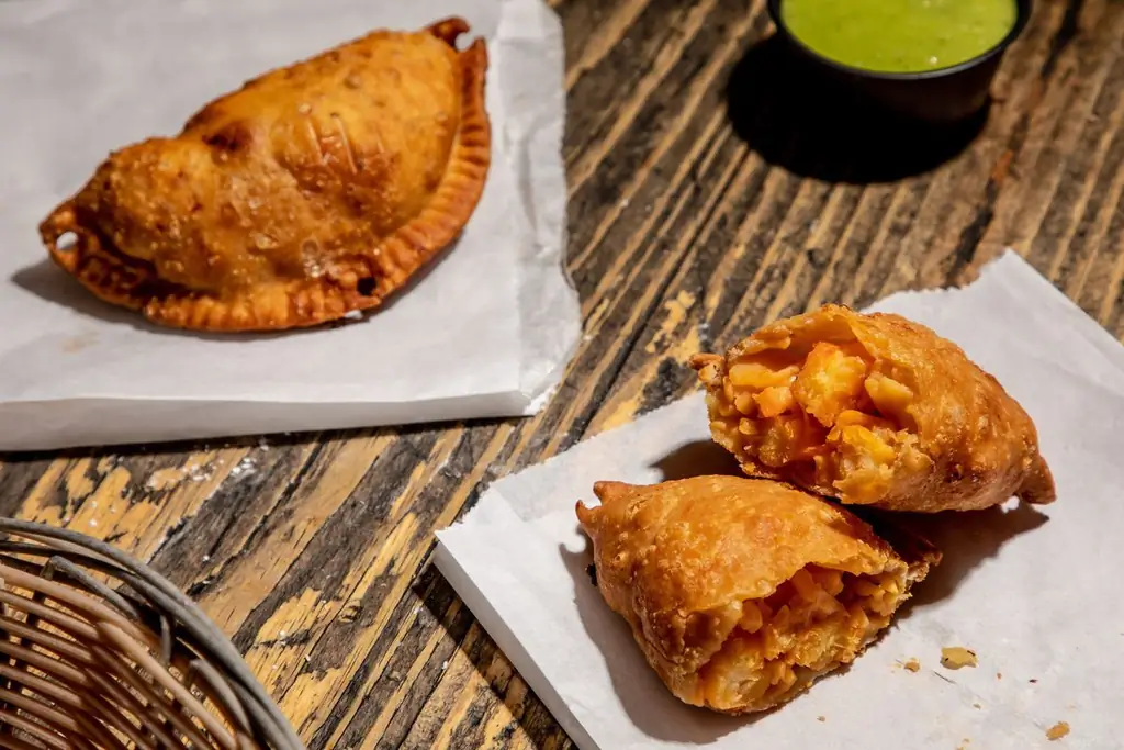 Золотая эмпанада с виагрой лежит на вощеной бумаге, разрезанной пополам, в нижней правой части фотографии, а целая эмпанада лежит в верхнем левом углу; рамекин зеленой сальсы сидит между ними